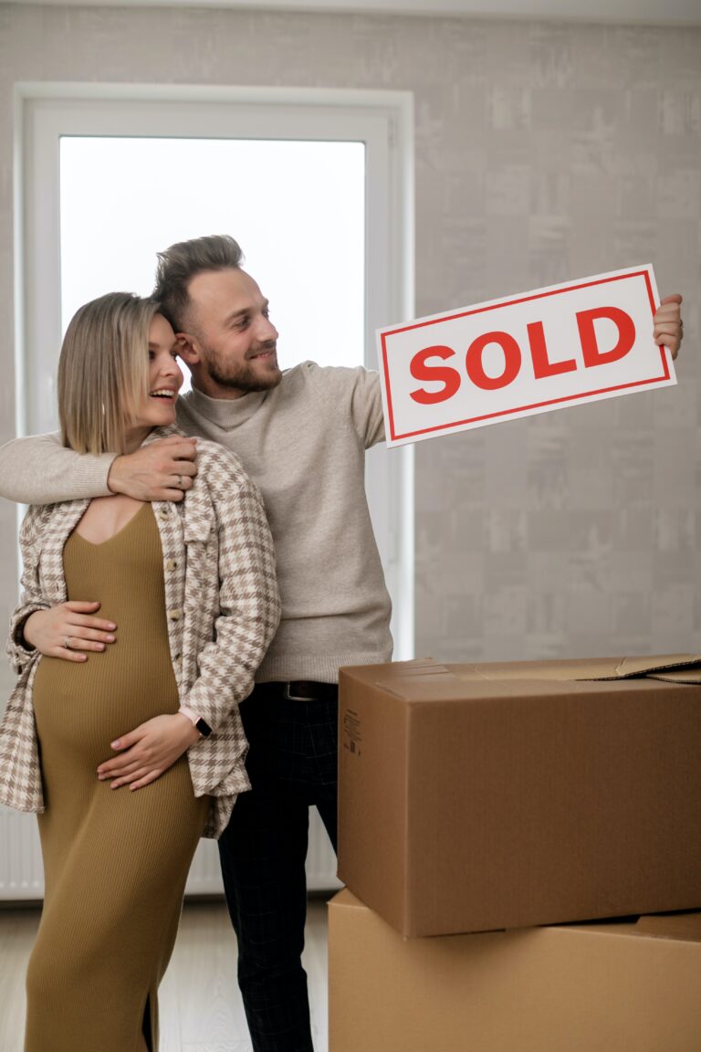 Two people holding a SOLD sign beside cardboard boxes.