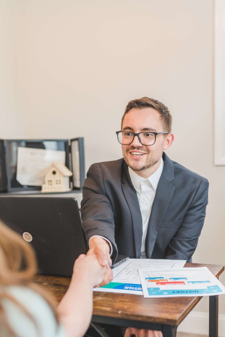 A person in a suit shaking hands across a desk with a laptop and property plans.